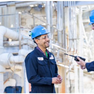 Two smiling people facing each other, wearing hard hats in an industrial area.