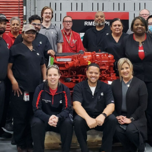 A team posed in a workshop with a large red engine.