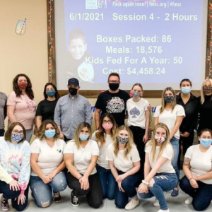 a group posed in front of a projected screen with stats of meals, cost, kids helped, etc