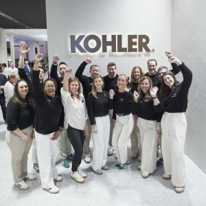 A team of people posed and cheering with arms up in front of a Kohler sign.