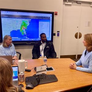 People meeting in a conference room. Digital screens with maps of the hurricane area displayed.