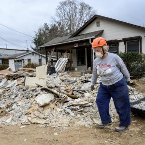 A volunteer in hard hat and respirator pulling a black sled full of rubble. A pile of rubble in front of the house behind them.