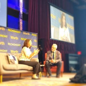 Heather Johnson and David Kirkpatrick seated on stage, a large screen behind them