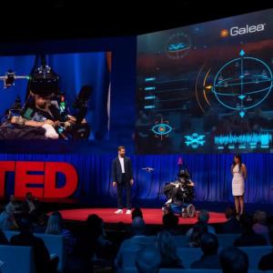 Three people on a stage "Ted" sign to the left, digital screens above.