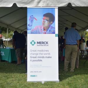 a poster board for Merck at the front of an outdoor tent, people mingling underneath or at tables