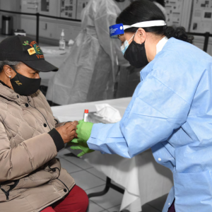 a person in protective medical apparel collects a sample in a test tube from another person seated in front of them