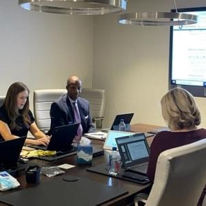 Three people seated at a conference table with laptops open. A digital display at the front.