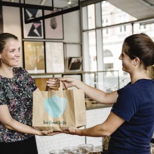 a person hands another a bag in a restaurant, the Too good to go logo on the bag