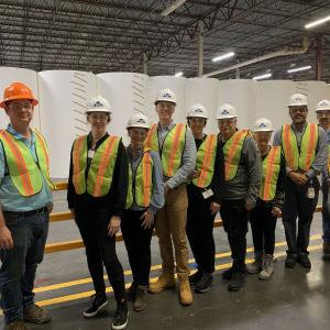 A line of people in high-vis vests and hard-hats posed in front of large rolls of paper in a large warehouse.