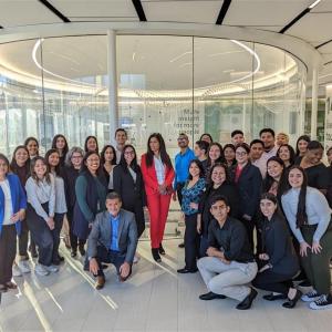 A group of people posed for the camera, some crouching in front, a large glass circular enclosure behind them in an office setting.