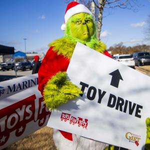 A person dressed as the Grinch holding a sign "Toy Drive" outside.