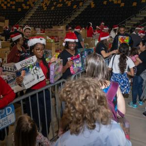 Volunteers in santa hats handing out toys to kids in a large auditorium.