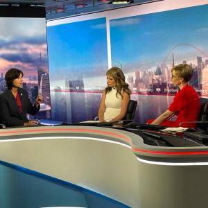 Tracey and two others seated behind a desk on a set.