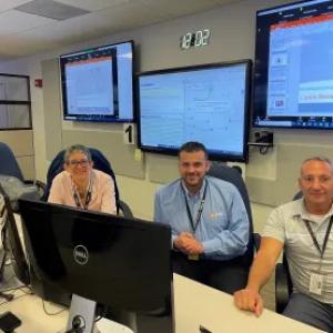 a group of three people, computer screen in front of them and many screens on the wall behind them