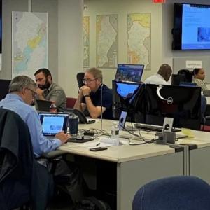 a large room with people seated at stationed tables, many screens on the wall