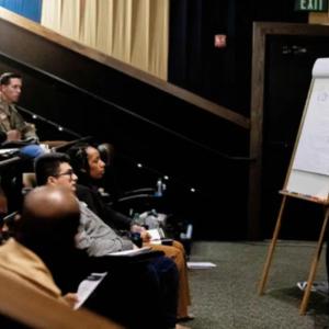 A person drawing on a paper board at the front of a room full of seated people. Some in military uniform.