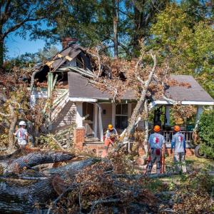 People in hardhats, some with chainsaws, watch as one cuts large limbs on a tree that has fallen on a small house.