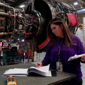 A person looking through a book, holding a clip board, an airplane engine suspended to their side.