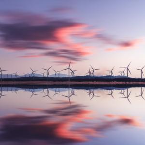 A group of wind turbines behind a body of water. The setting sun to the right.
