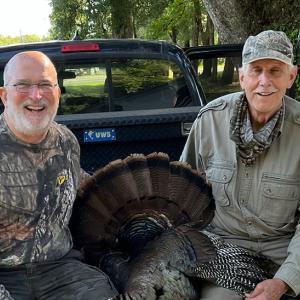 Two hunters with a turkey between them sitting in the back of a truck.
