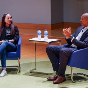 Two people seated on a lit stage, a small table between them. One speaking.