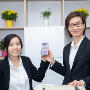 Two people holding the same cell phone. A bookcase behind them with a plant in each cube.