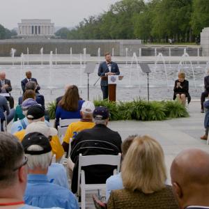 a person stands at a podium outside in front of a fountain memorial. There is a seated crowd and a panel of six others along side them