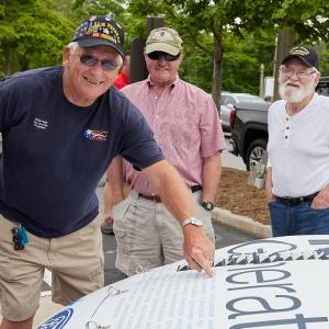 three veterans stand by a car, one points to a name on the hood