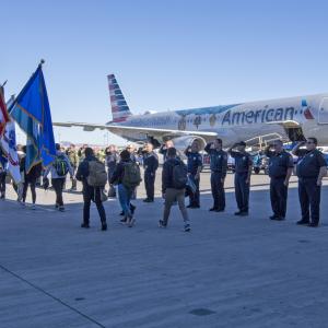On the tarmac, lines of uniformed officers holding flags and holding salute as other pass between them. A plane behind them.