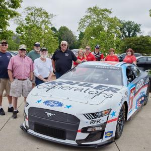 a group of people stand behind a race car, some wearing military service hats