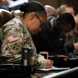 A person in military uniform seated next to others, writing in a notebook.