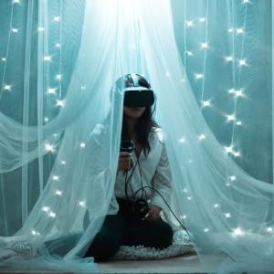 A person using a VR headset and controllers, sheer drapes and string lights around them.