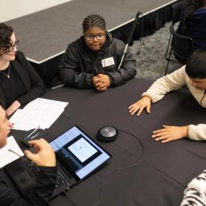 A table with five people seated. One with an open laptop pointed to the others.