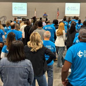 A person speaking on a stage to a group of volunteers and others standing in an open area.