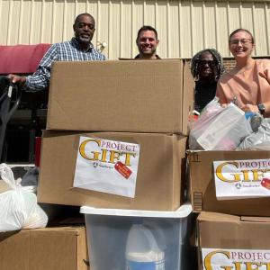 Four people posed behind boxes stacked on a walkway outside a building. "Project Gift" logos on some.