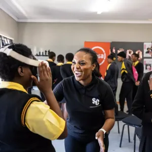 An adult smiles and assists a child with a vr headset. Others look on smiling.