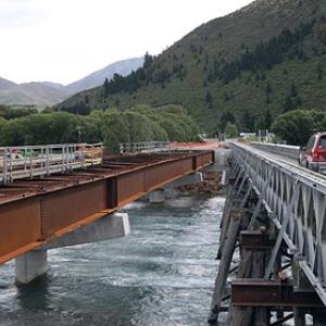 a car passes over a low bridge in mountainous terrain along side another bridge is under construction