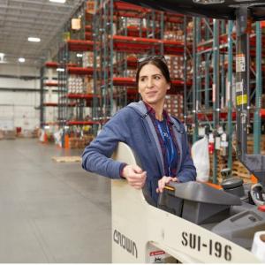 A woman in a warehouse stands in the operators seat of a forklift. She looks over her right shoulder and smiles with floor to ceiling shelves stacked with boxes behind her.