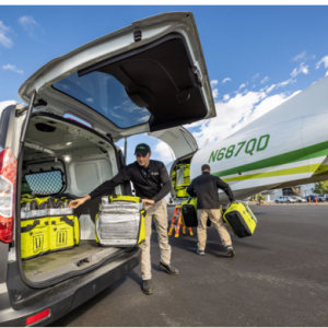 Two people unloading boxes from the back of a van, headed towards a small plane.