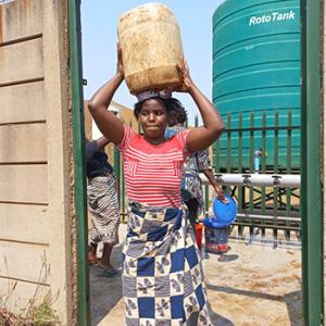 a person carrying a large jug on their head, a large tank behind them