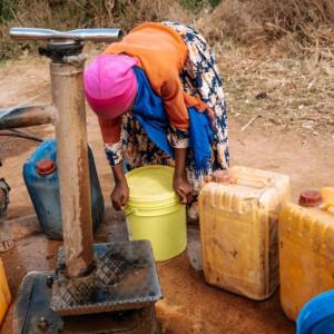 A person and two children at a well, pumping water into jugs.