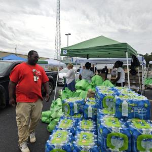 Stacks of wrapped water bottles outside, vehicles parked nearby