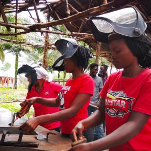 Three people each in a red shirt and welding masks, working at an outdoor table with welding tools and pieces of metal.