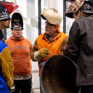 A teacher standing in a circle with students. All wearing welding helmets and protective clothing.