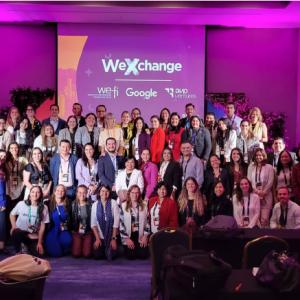 A large group posed in a room with purple lighting. A digital display "WeXchange" behind them.