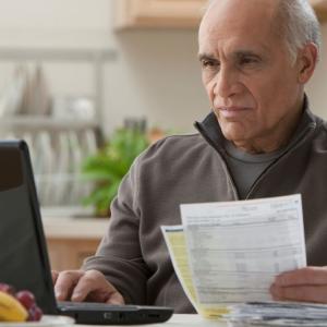 A person seated in front of a laptop computer in a home setting, holding papers in one hand.