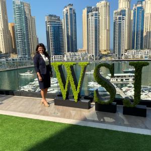 A person standing on a balcony next to green letters "WSJ" overlooking a body of water and skyscrapers.