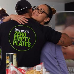 A person hugging someone with a t-shirt that reads: "One World, Zero Empty Plates"