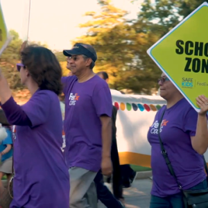 3 people in purple FedEx shirt, 2 of which are holding neon yellow "School Zone" signs