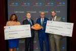 Four people posing with two oversized checks and a basketball